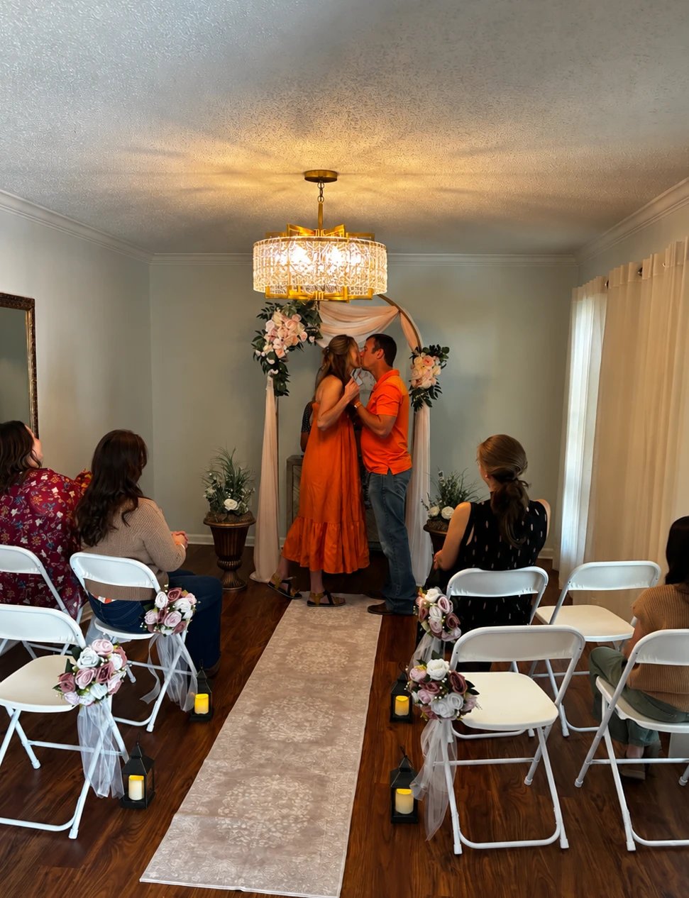 A couple sharing their first kiss under the blush draped floral arch and crystal chandelier, with guests seated on both sides of the aisle runner